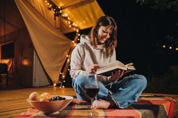 Smiling Woman freelancer drinking wine and read book sitting in cozy glamping tent in autumn evening. Luxury camping tent for outdoor holiday and vacation. Lifestyle concept