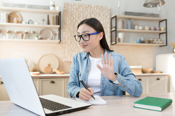 Remote work during quarantine. Young asian business woman freelancer working from home. Sitting at a table with a laptop, conducting an online meeting. conference. Says hello to the camera, writing.