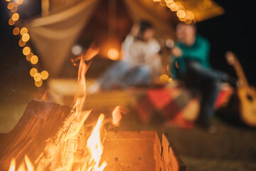 Happy couple relaxing in glamping on autumn evening, drinking wine and playing guitar near cozy bonfire. Luxury camping tent for outdoor recreation and recreation. Lifestyle concept