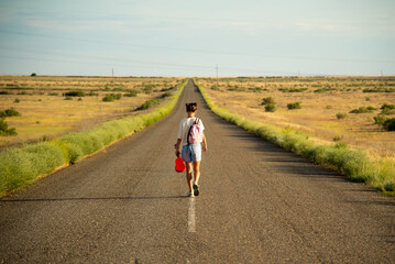 Obraz premium A girl walks along an empty road in the steppe. She has a ukulele in her hands and a backpack on her shoulders. It's summer and warm outside. Open road.