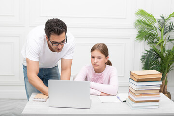 Father and daughter studying together online at home. He explaining lesson and she listening him carefully. Stack of books, laptop and exercise book on the desk. Online education concept