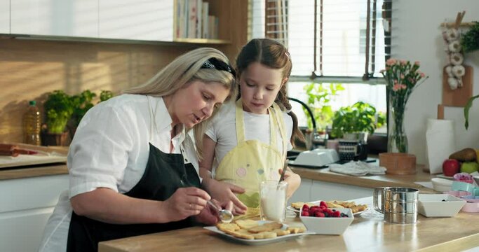 Elderly blonde granny with adorable preschooler granddaughter standing at wooden table in modern kitchen teaching cooking baking cookies in different shaped decorating with berries preparing surpirse.