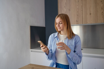 Attractive young woman texting on mobile phone, drinking water at kitchen. High quality photo