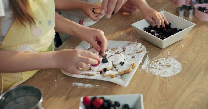 Close up shot selective focus on wooden surface with white plate. Kid's hands dressing decorating biscuits in different shapes with berries. happy family mother's day concept.