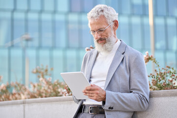 Gray haired old bearded stylish old mature adult professional business man, senior older businessman ceo wearing suit glasses holding tab using digital tablet fintech platform standing outdoors.