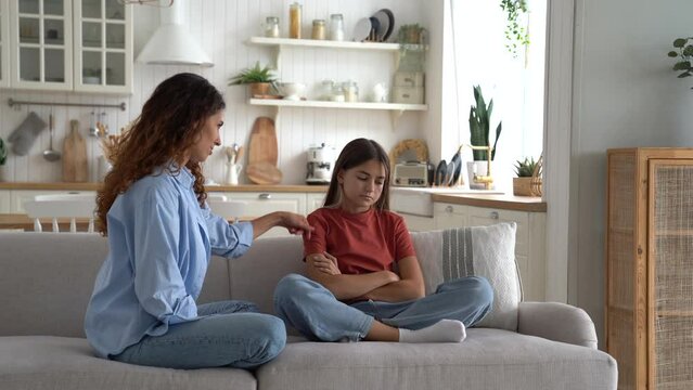 Good-natured mother tries to cheer up teenage girl and wants reconcile after punishing child for failing in school. Casual european woman tickling little daughter sits on sofa among home interior
