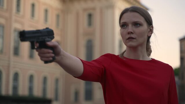 4k Young woman holds gun and shows shot at city street spbd. Close view of beautiful female is holding weapon in hand and imitating shot, standing against backdrop of stone building in town. One