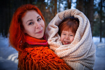 Mother with red hair and fluffy orange jacket and frozen little daughter in a warm blanket walking in winter, autumn or spring in forest. The concept of love and friendship of mom and girl