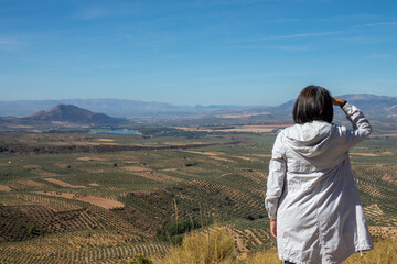 Fototapeta na wymiar Middle-aged Caucasian woman from the back admiring the views of the Andalusian landscape with olive groves and mountains from the Atalaya de Deifontes viewpoint (Granada, Spain), a sunny morning