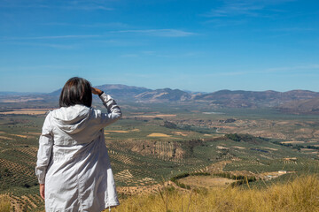 Middle-aged Caucasian woman from the back admiring the views of the Andalusian landscape with olive groves and mountains from the Atalaya de Deifontes viewpoint (Granada, Spain), a sunny morning