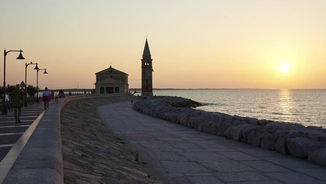 Time lapse of people stroll at dawn on the seafront of Caorle, in the background the sanctuary "Madonna dell'Angelo" built on a promontory surrounded by rocks.