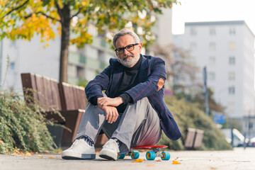 A smiling, elegant, bearded, grey-haired mature man with his glasses on, sitting on a red penny board in the city park, posing and looking at the camera. High quality photo