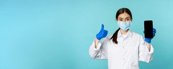 Portrait of doctor in medical face mask and gloves, showing mobile phone app, smartphone screen and...