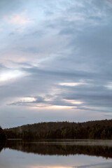 A tree-lined shoreline reflects with colorful clouds on a calm lake. Maple Leaf Lake, Algonquin Provincial Park, Ontario, Canada.