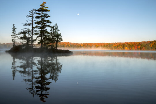 An island reflects on a calm lake with an autumn colored, tree lined shoreline and the moon in the background. Islet Lake, Algonquin Provincial Park, Ontario, Canada.