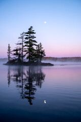An island's silhouette reflects on a calm lake with the moon above. Wonderful morning purple and mauve colors in the sky and water. Islet Lake, Alqonquin Provincial Park, Ontario, Canada.