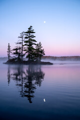 An island's silhouette reflects on a calm lake with the moon above. Wonderful morning purple and...