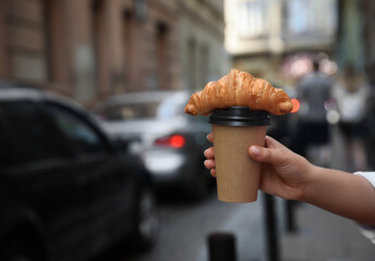Woman holding tasty croissant and cup of coffee on city street, closeup. Space for text