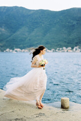 Fototapeta na wymiar Bride in a dress fluttering in the wind stands on the pier and looks at the sea