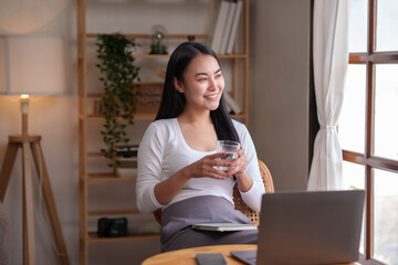 Beautiful Asian woman sitting and drinking water to relax from working on her laptop on the computer to reduce the fatigue caused by work at home.