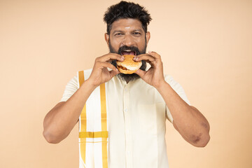 South indian man wearing traditional white dress eating burgers vada pav fast food snack isolated...