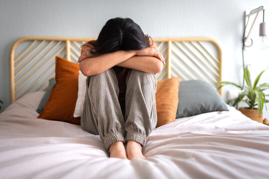 Front view of depressed and sad young woman sitting on the bed crying. Sadness and mental health.