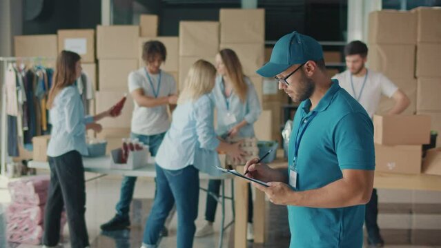 Male Volunteer Writing on Clipboard While Working in Food Bank Warehouse. Young Man Doing Inventory of Donated Goods in Volunteer Center While Group of Volunteers Sorting Humanitarian Aid