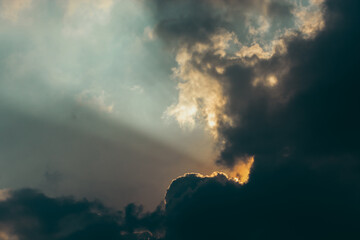 A view of a cloudy sky with the sun shining through the dark clouds. The light is casting rays down through the clouds.