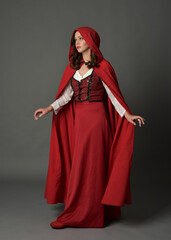 Full length portrait of beautiful brunette woman wearing red medieval fantasy costume with long skirt and flowing hooded cloak.
Standing pose with gestural hand poses, isolated on grey studio backgrou