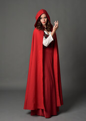 Full length portrait of beautiful brunette woman wearing red medieval fantasy costume with long skirt and flowing hooded cloak.
Standing pose with gestural hand poses, isolated on grey studio backgrou