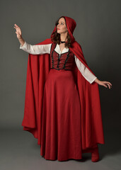 Full length portrait of beautiful brunette woman wearing red medieval fantasy costume with long skirt and flowing hooded cloak.
Standing pose with gestural hand poses, isolated on grey studio backgrou