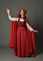 Obraz premium Full length portrait of beautiful brunette woman wearing red medieval fantasy costume with long skirt and flowing hooded cloak. Standing pose with gestural hand poses, isolated on grey studio backgrou