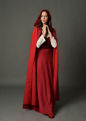 Fototapeta premium Full length portrait of beautiful brunette woman wearing red medieval fantasy costume with long skirt and flowing hooded cloak. Standing pose with gestural hand poses, isolated on grey studio backgrou