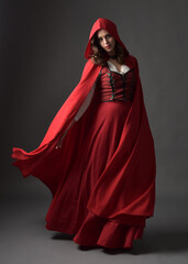 Naklejka premium Full length portrait of beautiful brunette woman wearing red medieval fantasy costume with long skirt and flowing hooded cloak. Standing pose with gestural hand poses, isolated on grey studio backgrou