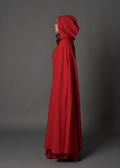 Naklejka premium Full length portrait of woman wearing red medieval fantasy costume, flowing hooded cloak. Standing pose side profile, gestural hand poses, walking away from camera isolated on grey studio background.