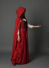 Full length portrait of woman wearing red medieval fantasy costume, flowing hooded cloak. Standing pose side profile, gestural hand poses, walking away from camera isolated on grey studio background.