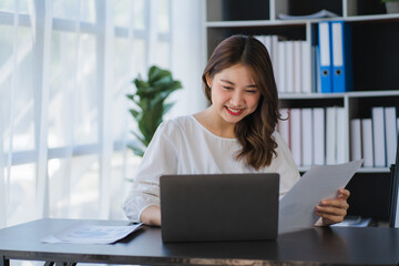 Fototapeta premium Beautiful Asian businesswoman working on a desk with a laptop with a smiling face while looking at the document received in the office.