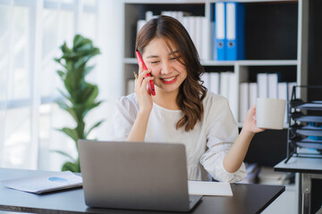 Asian businesswomen have the joy of talking on the phone with a laptop computer tablet on the office desk. doing accountant on a calculator to calculate business data documents.