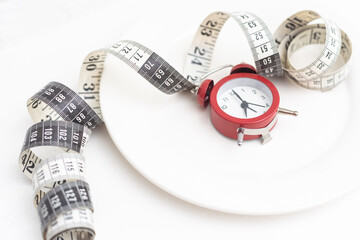 Time to lose weight concept. Red alarm clock with white plate and measuring tape close-up.