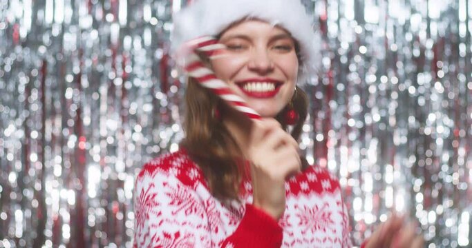 Smiling playful young woman in christmas sweater and hat posing and playing with candy cane on silver shiny backdrop foil curtain background