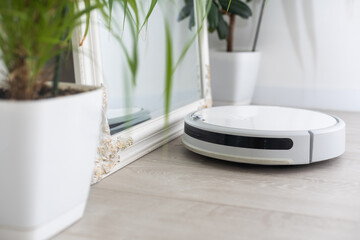 A white robot vacuum cleaner vacuums along the skirting Board near a large window through which the...