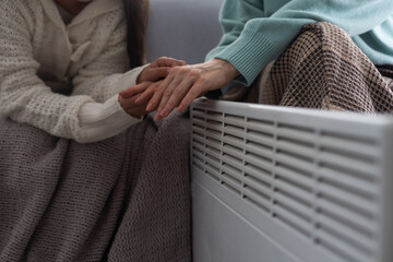 At the electric heater, people are warming themselves, covered with a warm blanket at home. hands of mother and daughter near the heater. Cold season and gas crisis in Europe