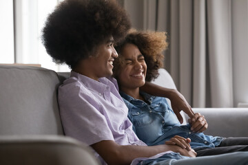 Laughing African teenager couple, girlfriend and boyfriend hugging on sofa enjoy pleasant conversation resting at home. First love, romantic relationship, friendship between girl and guy, family ties