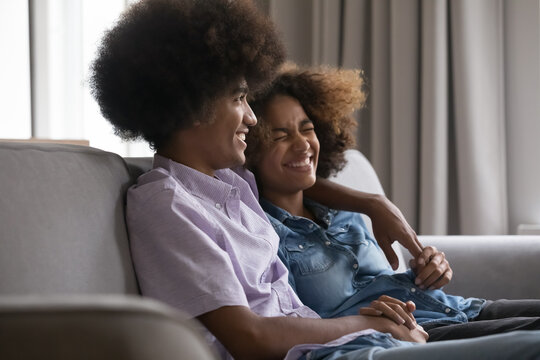 Laughing African teenager couple, girlfriend and boyfriend hugging on sofa enjoy pleasant conversation resting at home. First love, romantic relationship, friendship between girl and guy, family ties