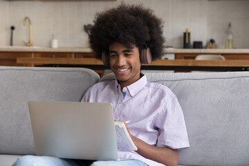 Teenager African student guy sit on sofa wear headphones take notes while studying remotely use...