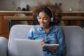 Fototapeta na wymiar African student girl sit on sofa wear headphones takes notes, studying remotely use laptop and video call application. Gen Z and modern tech usage, distancing learning at home, tuition, online class