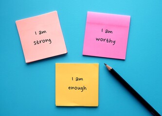 Three note paper on blue background with handwritten text I AM STRONG, I AM WORTHY, I AM ENOUGH -...