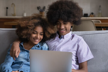 Fototapeta na wymiar Attractive African teenagers couple spend free time on internet using laptop, watching movie, enjoy new video vlog hugging resting seated on couch at home. Modern technology usage for fun and leisure