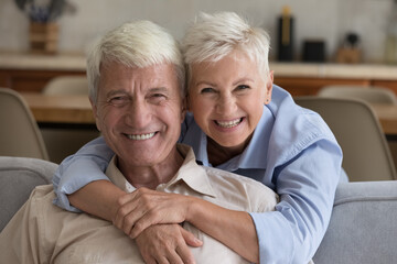 Grey-haired grandparents pose at home. Loving wife hugs husband seated on sofa staring at camera,...