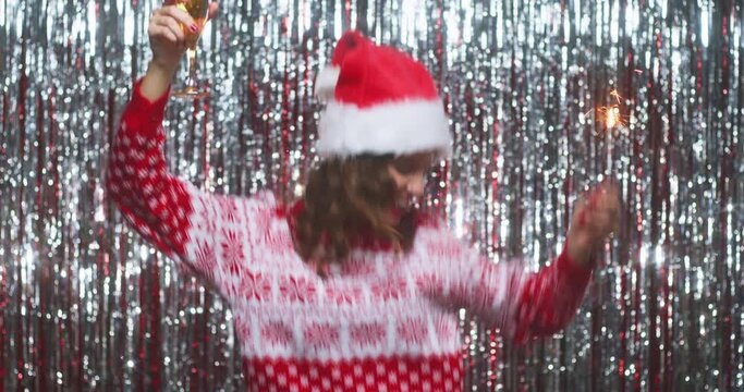 Woman raising glass of champagne and dancing on party with sparkler, dressed in christmas sweater and hat on silver foil curtain backdrop background
